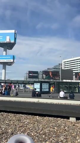 The video shows the exterior of Amsterdam Schiphol airport. A number of people are sitting on benches in front of the terminal building. A large white building with "SHERATON" written on it is visible in the background. There are several signs and advertisements visible, including one for "AMS Schiphol" and another for "DeGiro." There are cars and buses driving by the terminal, and people with luggage. The sky is blue with some clouds. The overall impression is that it is a busy airport with people waiting for transportation or relaxing outside. There are some people having food at a restaurant called "O Place".