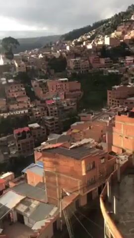 The video shows an aerial view of a densely packed neighborhood of buildings. Most of the buildings appear to be made of brick and have a reddish-brown color. They are nestled closely together on a hillside, with some greenery visible in between the structures. The sky overhead is overcast, suggesting it might be early morning or late afternoon. There is a bright flash of light in the middle of the video.