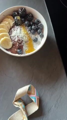 The video shows two smoothie bowls on a gray countertop. Each bowl contains a mixture of fruits, including sliced bananas, blueberries, and blackberries. One bowl also appears to have raspberries. The bowls are garnished with shredded coconut and a drizzle of honey or syrup. The lighting suggests an indoor setting, possibly a kitchen, with a window visible in the background. A white paper towel dispenser is also seen near the window. The camera moves slightly throughout the video, providing different angles of the smoothie bowls.