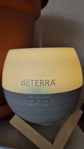 A close-up shot focuses on a white and gray essential oil diffuser. The diffuser is lit from within, casting a warm, yellow glow. The brand name "dōTERRA" is visible in gray text on the upper half of the diffuser. Below the brand name, there are three timer options for mist: 2H, 6H, and 12H, along with an icon representing a light. To the right of the mist options, there is a button labeled "LIGHT." The diffuser sits on a wooden stand composed of two pieces of wood angled in an X formation. In the background, a blurred brown object, likely a plant pot, is visible. The lighting in the scene is soft, with the diffuser being the primary light source.