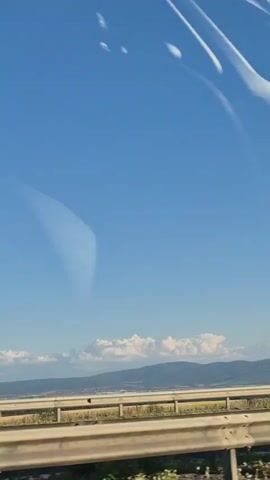 The video captures a view from a moving vehicle, likely a car, looking out at a clear blue sky. In the distance, there are rolling hills or mountains, with some white clouds scattered across the horizon. A highway or road barrier is visible in the foreground, suggesting the perspective is from a roadside. Power lines and a tall, lattice-style transmission tower are also prominent features of the landscape. The overall impression is of a bright, sunny day with a vast sky. Reflections on the window of the vehicle are also visible, adding a layer of visual complexity to the scene.