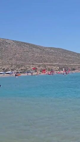 This video shows a bright, sunny day at a beach with clear blue skies and turquoise water. Numerous people are windsurfing in the water, with their colorful sails visible against the sea and the distant, dry, hilly coastline. Some people are also visible on the beach, and there appear to be some tents or structures set up along the shore. The water is calm and the scene depicts a popular water sports location.