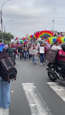 The video shows a parade with many people walking down a street. There are colorful balloons in the shape of rainbows and other decorations. Some people are holding signs and flags. The people are diverse in age and gender, and many are dressed in casual clothing. The overall atmosphere appears festive and celebratory. The video is shot from a slightly elevated perspective, looking down at the parade.