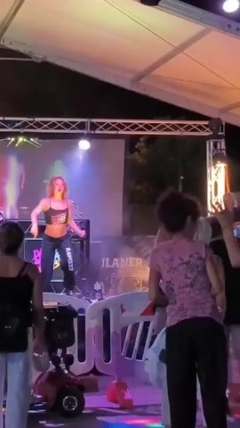 The video shows a woman dancing on a stage under a tent. She is wearing a black crop top and dark pants. The stage is lit with pink and purple lights. A large screen behind her displays silhouettes of dancers. There are speakers and other equipment on the stage. In front of the stage, a crowd of people is watching, some of whom are holding cameras or phones. Two mobility scooters are parked in front of the stage barrier. The environment appears to be an outdoor event at night.