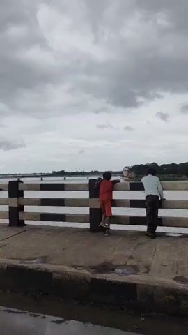 The video shows a view from a bridge with a railing. The railing has alternating black and white bars. Beyond the railing, there is a body of water. In the distance, there is a shoreline with some vegetation. The sky is overcast with gray clouds.