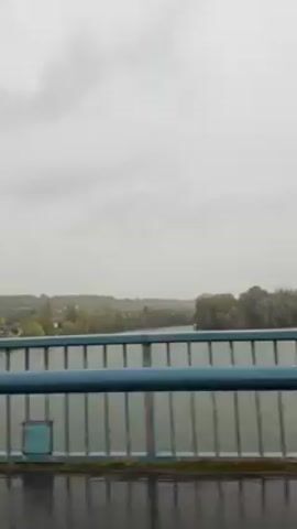 The video shows a view over a river from a bridge on a cloudy day. The bridge has blue railings. The river is wide and calm, and there is some vegetation on the banks in the distance. It appears to be raining.