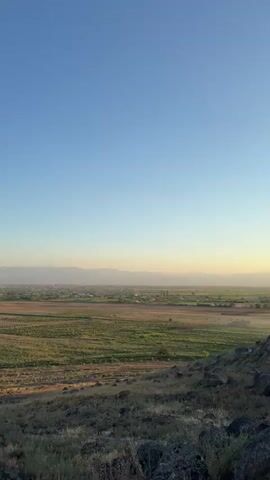 This vertical video captures a panoramic view of a rural landscape in Nor Armavir, Armenia, during what appears to be late afternoon or early evening. The sky is a clear, pale blue, transitioning to a softer hue closer to the horizon.

In the foreground, a rocky, arid slope covered with dry grasses and sparse bushes creates a textured foreground. Beyond this, a vast agricultural field dominates the mid-ground, appearing to be harvested or in a dormant state, with rows of tilled earth or stubble stretching across the expanse. A few scattered trees and a thin line of vegetation mark the edges of the field.

Further in the distance, a town or village is nestled between the fields and the mountains. Buildings with light-colored roofs are visible, suggesting a rural settlement. The primary focal point of the distant scene is a majestic, snow-capped mountain range, likely Mount Ararat, which rises prominently against the horizon, its peak illuminated by the setting sun. The mountains on either side appear as softer, hazy silhouettes.

The overall mood is serene and tranquil, emphasizing the vastness of the landscape and the imposing presence of the mountains. The warm, golden light cast on the fields and the distant mountain suggests a peaceful end to the day. There is no discernible activity or people depicted in the video. No visible text is present.