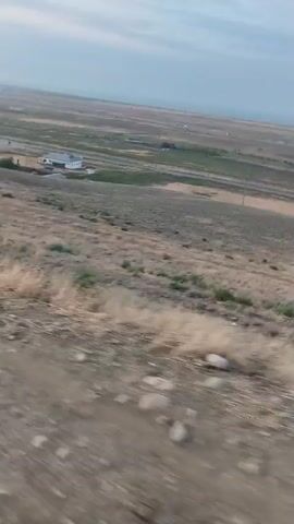 A video captures an outdoor scene from a moving vehicle, likely a car, on a dirt road. The camera perspective is low, looking out and slightly downwards. The terrain is hilly and covered with dry grass and scattered rocks. In the middle distance, a white house with a dark roof is visible, situated on a gentle slope. Beyond the house, the land stretches out into a vast, open plain under a hazy sky. The sky appears overcast or dusty, with a pale, diffused light. The video is shaky, indicating movement and possibly a bumpy ride. The overall impression is one of a rural, undeveloped landscape.
