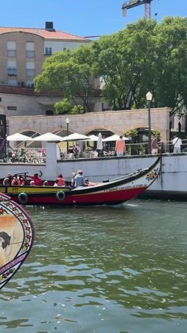 A red and yellow boat carrying approximately ten people is moving on a body of water. The boat has a distinctive curved bow and stern, with green and red fenders along its sides. Some passengers are looking towards the camera or at each other, while others appear to be enjoying the scenery. In the foreground, another boat with a decorative circular element featuring a painted image is partially visible. In the background, there is a stone embankment with white umbrellas and tables, suggesting an outdoor seating area for a restaurant or cafe. Behind that, a building with several stories and a red-tiled roof is visible, along with lush green trees. A construction crane can be seen in the upper right portion of the frame. The sky is clear and blue, indicating a sunny day. The water is calm and reflects the surrounding environment.