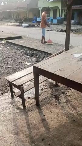 A person is sweeping the ground with a broom. The person is wearing an orange shirt and blue shorts. The person is standing in front of a wooden table and bench. There are houses in the background. The ground is dirt and gravel. The sun is shining.