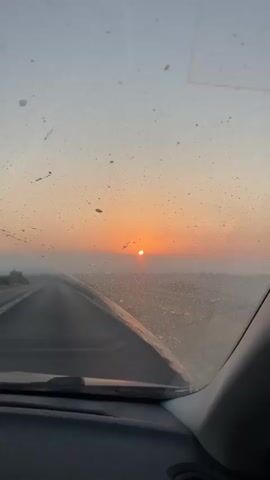 The video shows a view from inside a car as it travels on a dusty road during sunset. The sun is low on the horizon, casting an orange glow across the sky. The car's windshield is dirty, with specks of dust obscuring parts of the view. The road ahead appears to be in a desert or arid landscape. In some frames, mountains or hills can be seen in the distance, and the sky transitions from orange and pink near the horizon to a pale blue overhead. A guardrail is visible along the side of the road in some shots. The interior of the car, including the dashboard and a side mirror, is partially visible in some frames. The overall impression is of a journey through a remote, scenic location at dusk.