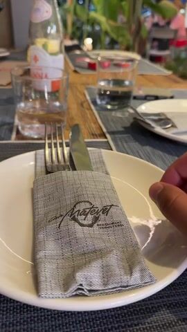 The video shows a table setting at a restaurant. In the foreground, a white plate has a gray napkin with a logo that reads "Matevet Restaurant & Cafe". A fork and knife are placed inside the folded napkin. To the left of the plate is a glass of water and a bottle of Acqua Panna mineral water. To the right of the plate is another glass of water. In the background, there are more tables with place settings, plants, and people dining. Some people are walking past the table. The lighting suggests it is evening.