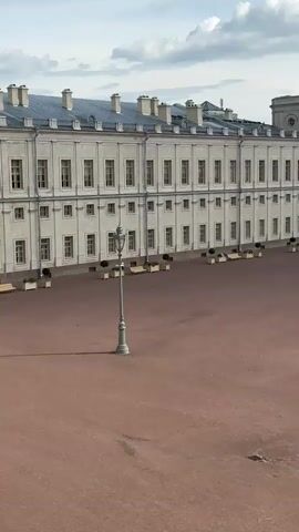 The video captures a wide shot of a large, open, reddish-brown courtyard in front of a grand, pale yellow building with many windows. A single, tall, ornate lamp post stands in the center of the courtyard. Along the side of the building, there are planters with green foliage and some benches. In the distance, a line of trees forms a backdrop, and beyond the trees, there are parked cars, more buildings, and a road with some traffic, including a white bus. Two people are walking across the courtyard in the middle distance. The sky is blue with scattered white clouds. The camera pans slowly to the right, revealing more of the courtyard and the surrounding landscape. Another person is seen walking in the far distance. The overall impression is of a historic and spacious outdoor area.