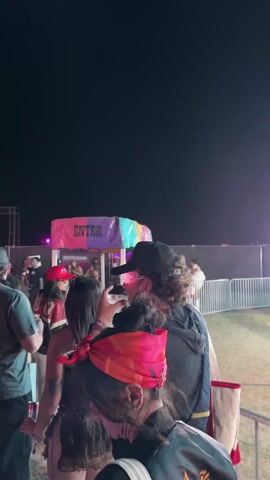 The video shows a fair at night. In the foreground, there are a few people, and then a metal fence. Beyond the fence is a fairground ride called "Tornado" which is brightly lit with white lights. People are sitting in the ride. There is a worker standing near the ride. In the background, there are some food stands and more people. The sky is black.