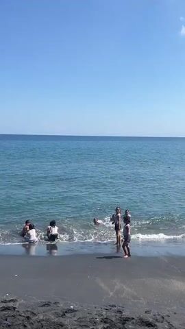A vertical video shows a group of children playing in the shallow ocean water near a dark sand beach. The sky is clear and blue. In the foreground, a boy wearing a white t-shirt and black shorts stands on the beach, facing the ocean and raising his right arm. To his left, in the water, a group of several children are splashing and playing. One child in the middle right of the frame is bent over in the water. A small kite is visible in the lower right corner of the frame. The camera is still.