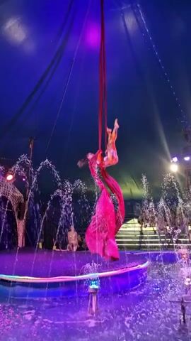 A person performs aerial silks in a circus tent. The person is suspended by two red silk ropes and is wearing a sparkly costume. Water jets spray in the background, creating a misty atmosphere. The tent is illuminated with blue and purple lights.
