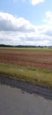 A dirt road borders a large, recently plowed brown field under a bright blue sky with fluffy white clouds. A few trees are visible in the distance on the horizon, and a thin line of green grass runs along the edge of the road. Two tractors are plowing the field in the distance, kicking up dust as they work. The video is a static shot from a moving vehicle, capturing the agricultural landscape.