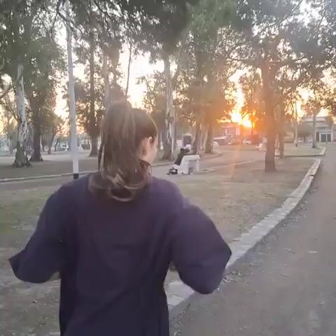 A person with long brown hair tied in a ponytail, wearing a dark purple sweatshirt and black leggings, runs along a dirt path. The path is bordered by a low stone wall on the right. The background features a park-like setting with trees and grass. The sun is setting, casting a warm orange glow through the trees. The camera is positioned behind the runner, following their movement.