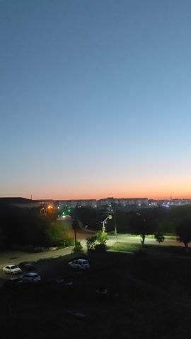 The video captures an urban landscape during twilight. A clear sky transitions from a deep blue on the left to a soft orange and pink hue on the right, indicating the setting or rising sun. Below the sky, a city skyline is visible with buildings, some illuminated, and streetlights casting pools of light on a road. Trees and greenery are interspersed within the urban environment. In the foreground and to the right, the scene darkens considerably, suggesting closer proximity to the viewer or less ambient light. Several parked cars are discernible in the darkened area. The overall impression is a peaceful, transitional moment between day and night in a city.