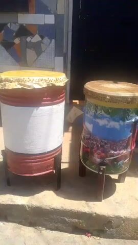 The video shows two drums standing on stands outside. The drum on the left is white and red, and the drum on the right is decorated with a colorful mural. In the background, there is a building with a tiled wall and a dark doorway. Two people are visible in the video: a young woman standing to the left of the drums, and the legs of another person visible on the right side of the screen, standing behind the drums. The scene appears to be in an outdoor, possibly urban, environment, with sunlight casting shadows. The video is static, with no camera movement.
