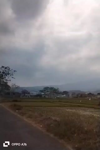 The video shows a wide shot of a rural landscape with rice paddies in the foreground and a village nestled in front of rolling hills and a mountain in the background. The sky is overcast with thick clouds. The rice paddies appear to be recently flooded or prepared for planting, with water reflecting the sky. The village consists of scattered houses with red tile roofs. A road is visible on the left side of the frame. The video is taken from a moving perspective, suggesting it was filmed from a vehicle. The OPPO A76 watermark is visible in the lower left corner. The overall mood is calm and serene, typical of a rural setting under cloudy skies.