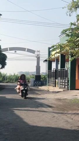 A woman wearing a headscarf and a wide-brimmed hat rides a motorcycle down a dirt road. She is carrying several bags, which appear to be plastic shopping bags, on the sides of her motorcycle. In the background, another person on a motorcycle is visible, as well as a building with a green and yellow facade. The sky is clear and blue, and the sun is shining. The overall scene suggests a rural or suburban setting in a warm climate.