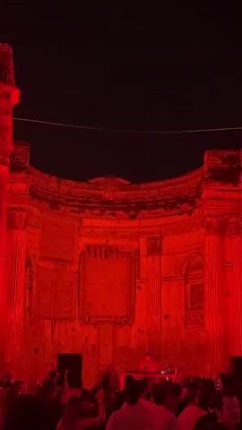 A crowd of people are gathered at an outdoor event. A DJ is set up on a platform in front of an ancient brick structure that is illuminated with red light. A projection of an eye appears on the structure, changing in appearance throughout the video. The overall mood is energetic and festive.
