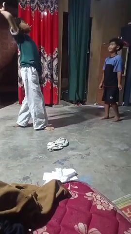Two young boys are playing indoors. The boy on the left, wearing a green shirt and white pants, is in motion, appearing to toss a red balloon upwards. The boy on the right, wearing a blue and black shirt and black shorts, stands with his arms raised, ready to catch the balloon. The room has a concrete floor and is sparsely furnished, with red and green curtains visible on the walls, a two-door cabinet, and a motorcycle parked in the background. Several items of clothing are strewn on the floor in the foreground.