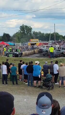 The video shows a demolition derby event. Several cars, damaged and with debris, are positioned in an arena. A crowd of spectators stands behind a barrier, watching the event. Several individuals in safety vests and hard hats are present in the arena, seemingly overseeing the event. The sky is cloudy with patches of blue. The environment appears to be an outdoor fairground or fair.