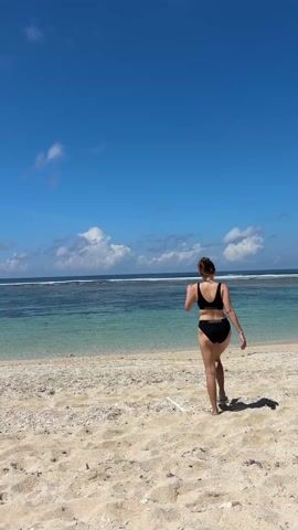 A woman in a black bikini walks away from the camera on a sandy beach toward a turquoise ocean under a bright blue sky. The beach appears sparsely populated, with sand and some pebbles or small stones. The ocean has some visible waves in the distance and appears calm near the shore. The sky is clear with a few puffy white clouds.
