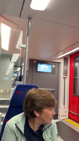The video shows the interior of a train carriage. In the foreground, a woman with short brown hair wearing a light green jacket is visible, slightly out of focus. The seats are upholstered in blue fabric with a red pattern. The carriage appears to be moving, as indicated by the blurriness of the view through the windows. Further down the aisle, more blue seats are visible. The ceiling is equipped with bright fluorescent lighting. A red door is visible on the right side of the frame, and a digital display screen is mounted on the wall to the left of the door. The overall impression is of a clean and modern train interior.