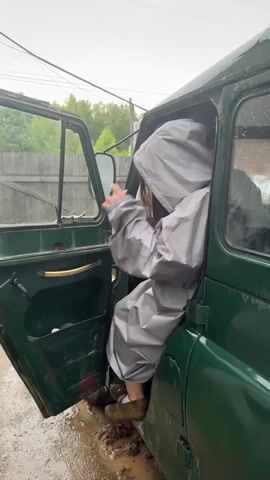A person is sitting in the driver's seat of a green vehicle. The door is open, and the person is wearing a grey hooded rain suit and dark sunglasses. The ground outside the vehicle is muddy, and there are visible puddles. The person appears to be either getting into or out of the vehicle, and they are looking at the camera. The background shows a wooden fence and some trees. The weather appears to be overcast, possibly raining. The person speaks in Russian, saying "Where are we going?" and "We're going into the forest."