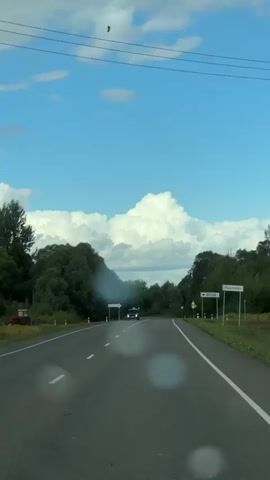 A vehicle is driving on a two-lane road. The road is paved and has white lane markings. There are trees on both sides of the road, and a cloudy blue sky above. A black car is driving in the same direction as the camera vehicle, and it appears to be emitting smoke from its exhaust. There are road signs visible on the right side of the road. The video is taken from the perspective of the driver or passenger of a vehicle.