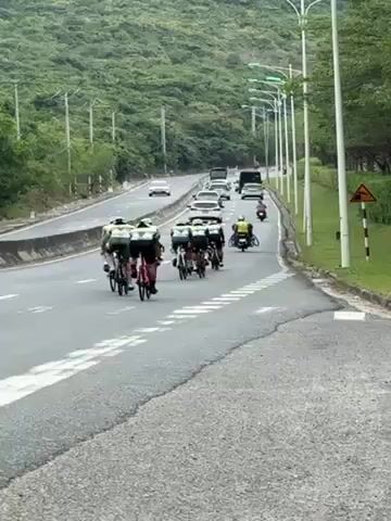 A group of cyclists ride on a road, followed by a motorcyclist. The road has multiple lanes and traffic. There's a line of streetlights along one side, and a green, hilly area is visible in the background. The motorcycle is carrying an additional bicycle on its back. The cyclists are wearing matching uniforms.