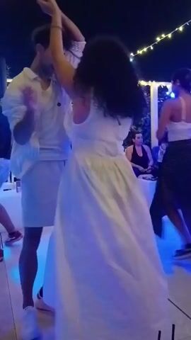 A group of people are dancing outdoors at night. In the foreground, a man and woman in white are dancing close together. The woman is wearing a long white dress. Behind them, a woman in a polka dot skirt and white top is also dancing. Further back, several people are seated, watching the dancers. The scene is lit by string lights and colored lights on the dance floor.