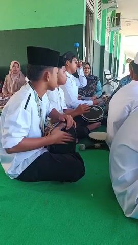 The video shows a group of people sitting on a green carpet, likely indoors or under a covered area. In the foreground, three young males wearing white shirts, black pants, and black skullcaps are playing hand drums. The person on the far left is playing a large, dark-colored drum with both hands. To their right, another young male is also playing a similar drum. The third young male further to the right is playing a smaller, lighter-colored drum with one hand while holding a stick in the other, as if to strike it or another instrument. In the background, to the left, a woman wearing a hijab is seated. Behind the drummers, more people are visible, including a woman with a headscarf and another person. A microphone is placed on the carpet in front of the drummers. The environment appears to be a building with a green wall and windows. Sunlight is visible, suggesting it's daytime. The overall scene depicts a musical performance, likely religious or cultural, given the attire and instruments.