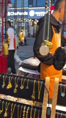 The video captures a street market scene in Linghe, China, likely in the evening as indicated by the artificial lighting and the dim sky. A vendor, a man wearing an orange apron over a dark jacket, is at his stall, which is laden with various jewelry items. He is holding a smartphone and appears to be engaged in a conversation or checking something on it.

The stall displays a wide assortment of necklaces, bracelets, and pendants. Many of the jewelry pieces are gold-colored, featuring intricate designs, including cloverleaf shapes, butterflies, and larger decorative pendants. Some appear to be made of jade or similar stones. The items are neatly arranged on black display stands and tables, creating a visually appealing presentation.

In the background, other people are seen browsing or walking past the market stalls. The setting suggests a bustling commercial area with shops lining the street, one of which has a sign with "ChaPanda" visible. Other signs in Chinese characters are also present, indicating the location is likely in a Chinese-speaking region. The overall atmosphere seems lively, with the vendor focused on his business and customers moving through the market. The weather appears to be fair, with no immediate signs of rain or strong wind.