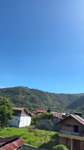 This is a vertical video of a village near a lake. The video begins with a view of houses with red roofs and green hills in the background. The sun is visible in the sky. As the video progresses, the camera pans to the right, revealing a wider view of the village and the lake. There are several houses with red roofs, some with metal roofs, and some with white walls. There are also several boats on the lake, some of which appear to be floating houses. The lake is surrounded by green hills and mountains. The sky is blue with some clouds.