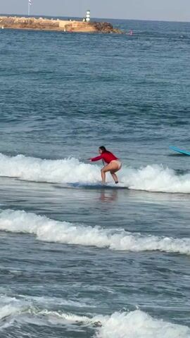 A person in a red top and light-colored bottoms is surfing on a blue surfboard in the ocean. The person is riding a wave, and their body is positioned in a crouched stance, with arms extended for balance. The water is blue, and there are white crests on the waves. In the background, there is a breakwater made of rocks, with a lighthouse or tower on it, and a few small white structures. The sky is clear and blue.