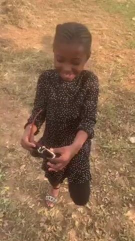 A young girl, around preschool age, smiles at the camera. She has dark skin, dark eyes, and her hair is braided into neat rows. She's wearing a black, long-sleeved dress with small white polka dots, black leggings, and sandals. She is holding something small and white in her hands. The background is a grassy area with scattered brown leaves. A person wearing black pants and black shoes walks into the frame and the girl reaches for them.