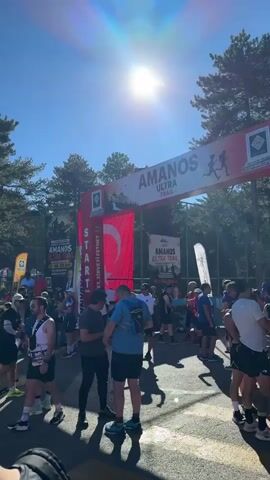 The video shows a crowd of people gathered outdoors under a clear blue sky, with a bright sun casting lens flares. They appear to be participating in a race, possibly a trail run, as indicated by banners that read "AMANOS ULTRA TRAIL" and "START." Many individuals are wearing athletic clothing and some have race bibs. There are tents and flags set up, suggesting an organized event. The environment is natural, with trees visible in the background, indicating a park or mountainous area. People are standing, talking, and some are stretching or adjusting their gear. The overall atmosphere seems to be one of anticipation and preparation before a race begins.