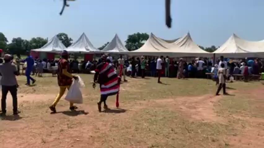 The video shows an outdoor event on a sunny day, with several white tents set up on a grassy field. Many people are walking around, some dressed in traditional clothing. There are also a few hand-pulled carts visible. The atmosphere appears to be social and celebratory.