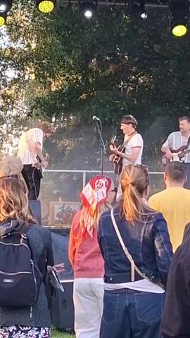 A band is performing on an outdoor stage. The band members are playing guitars and appear to be singing. The stage is lit by spotlights, and there is smoke or fog on the stage. A crowd of people are watching the performance from the front. The weather appears to be good, with sunlight visible through the trees behind the stage.