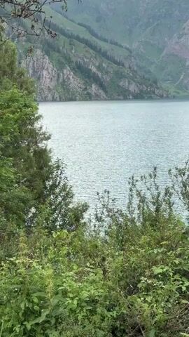 The video shows a serene mountain lake surrounded by lush green trees and steep, rocky mountains. The water is a light blue color and appears calm with a slight ripple. The mountains in the background are covered in green vegetation and have rocky outcrops. The sky is overcast with some clouds visible. The camera pans slightly to the right, revealing more of the lake and mountains. The scene is tranquil and picturesque.