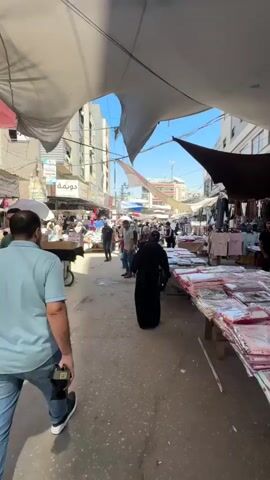 A first-person perspective video shows a bustling marketplace with a narrow walkway. The camera is positioned behind two people walking down the path. To the right, market stalls display clothing and other goods, some folded on tables and others hanging from racks. A torn fabric canopy stretches across the top of the scene, providing partial shade. Buildings line both sides of the walkway, with windows and signs visible. Several other people are walking in various directions through the market, some appearing to be shoppers and others vendors. The overall atmosphere suggests a lively and crowded commercial area under a clear blue sky.