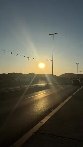 The video shows a sunset viewed from a road. The sun is setting behind a distant mountain range, casting a golden glow across the sky and the road. A tall lamppost stands on the right side of the road, and a string of small flags is strung across the top left of the frame. A white car is visible on the road in the distance. The road itself is paved and has lane markings. The sky is clear and a pale blue. The video pans slightly to the right, showing more of the road and the distant landscape.
