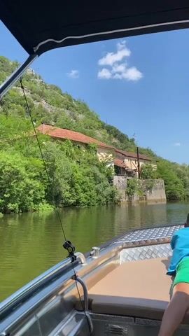 This is a first-person perspective video taken from a moving boat on a river. The boat has a metal railing in the foreground and a black canopy above. The river water is a murky green color. On the right side of the frame, a portion of a person wearing a green shirt is visible. On the left bank of the river, a steep, rocky hillside covered in green vegetation is seen. In the middle ground, nestled among the trees and extending up the hillside, are several old, dilapidated buildings with red tile roofs. These buildings appear to be abandoned, with visible structural damage and overgrowth. The sky is blue with a few white clouds. The boat is moving forward, causing the scenery to glide by.