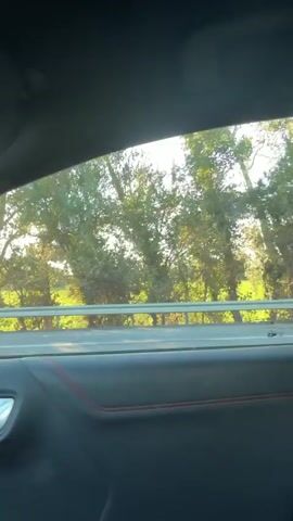 The video is taken from inside a moving vehicle looking out the window. In the distance, there is a large red fire truck with yellow accents and a person in a high-visibility vest on its back. Following the fire truck is a red pickup truck with flashing blue lights. Further down the road, two people in high-visibility vests are walking along the median barrier. The scene appears to be a highway with trees and vegetation alongside.