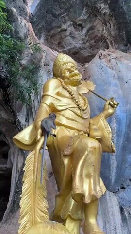A monkey sits on the arm of a large, golden statue of a man. The statue wears a golden headdress and beads, and holds a golden staff with a feather-like object at the top. The statue is seated in front of a rocky, cliff-like environment with some greenery visible. The monkey is facing the statue and appears to be looking at it.