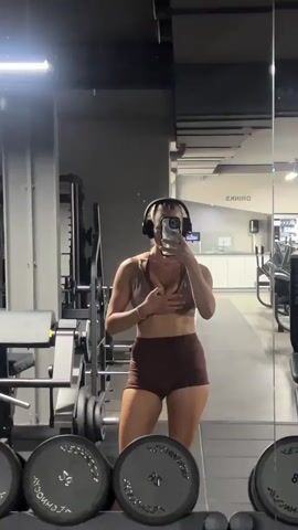 A woman wearing headphones and a brown athletic set takes a mirror selfie in a gym. She is standing in front of a rack of weight plates, with gym equipment visible in the background. She adjusts her top and flexes her arm.